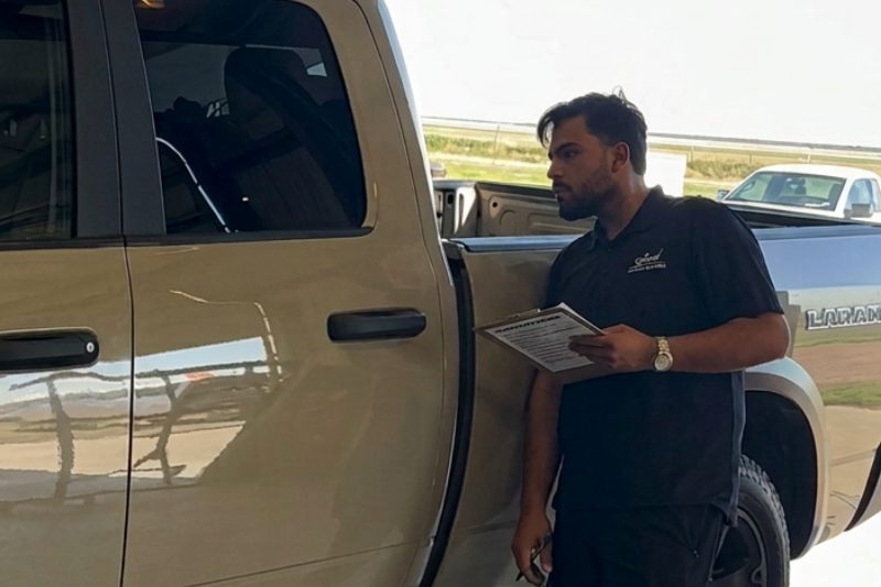 A technician beside a tan pickup, filling out an inspection sheet on a clipboard