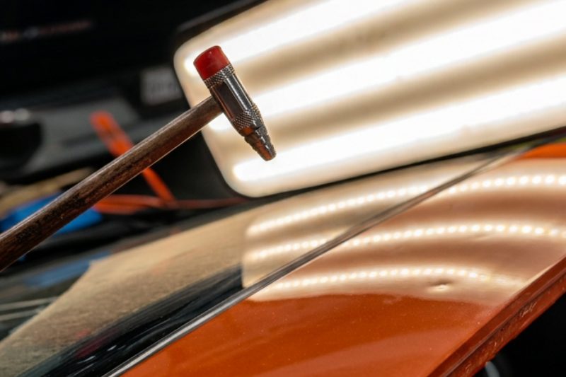 A dent tool tapping down a repaired panel on an orange roof under a striped light