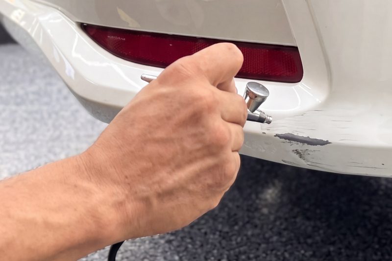 A hand holding a small spray gun over a scuffed white bumper