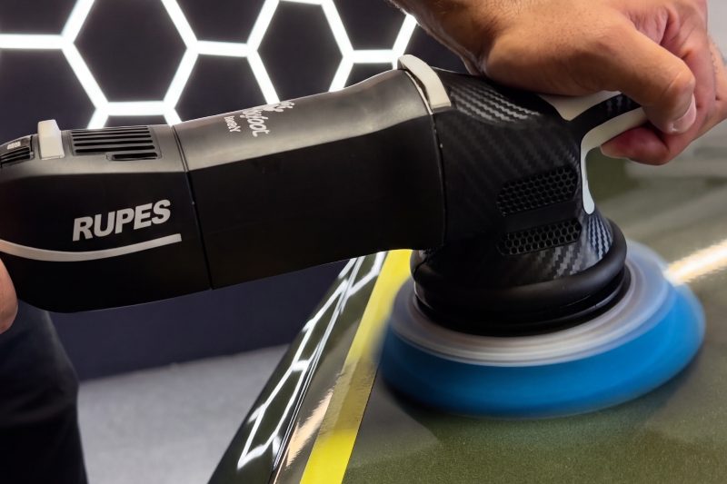 A hand guiding a dual-action polisher with a blue pad across a masked panel