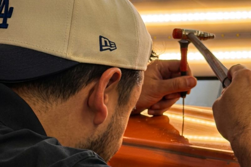 A technician leaning close over an orange panel, tapping a dent down with a hand tool